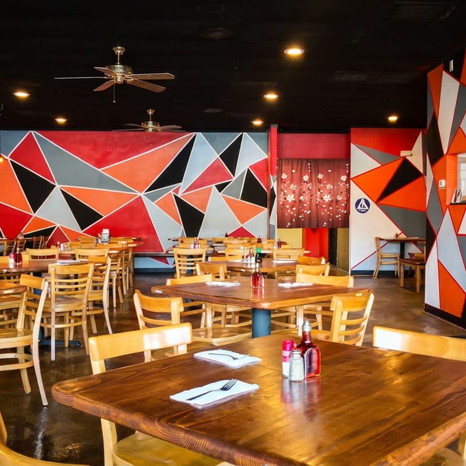 Empty restaurant dining room with wooden tables, chairs, place settings, ceiling fans, televisions, and vibrant geometric red, orange, black, and white walls.