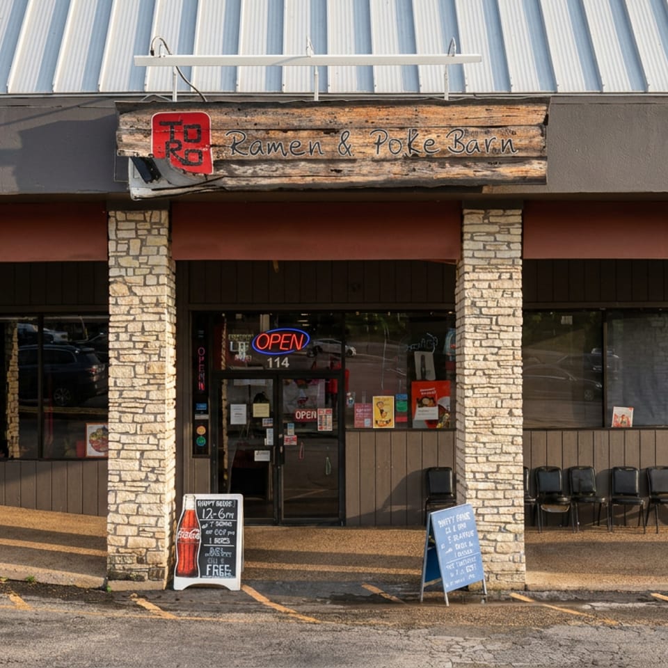 Exterior storefront of Ramen & Poke Barn with stone columns, illuminated open sign, windows, entrance, and sidewalk boards.