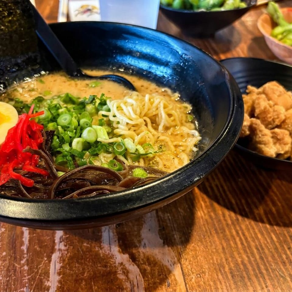 Ramen bowl topped with egg, green onions, seaweed, red ginger, and mushrooms beside fried chicken on a wooden table.