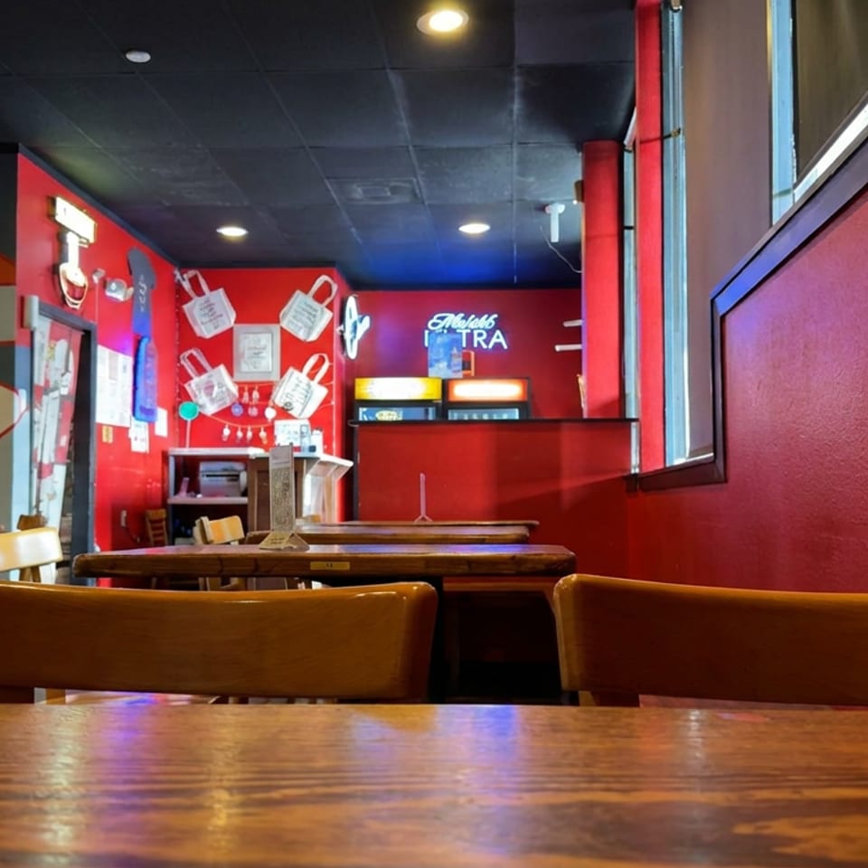 Colorful casual restaurant interior with wooden tables, chairs, framed posters, red walls, and a brightly lit service counter.