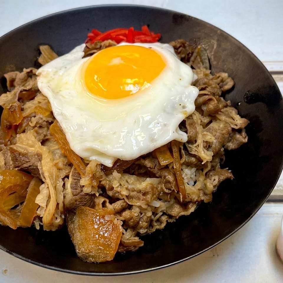 Black plate of stir-fried meat and vegetables over rice, topped with a sunny-side-up egg, with chili seasoning on the side.