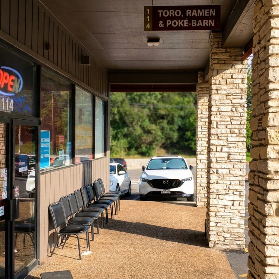 Restaurant storefront with neon open sign, menu board, outdoor waiting chairs, stone columns, and sign for Toro Ramen & Poke-Barn.